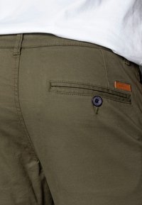 Olivegröna byxor i bomullsblandning, med en ficka med knappstängning, subtil textur och en läderetikett med varumärket.