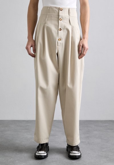 Pantalons beiges taille haute à jambe large, avec une fermeture à bouton à l'avant, des plis et une texture lisse. Portés avec des chaussures noires.