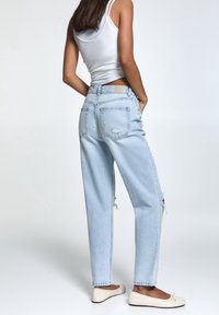 Donna con jeans strappati di colore azzurro chiaro, top bianco e ballerine color crema, in piedi con le mani nelle tasche su uno sfondo neutro.