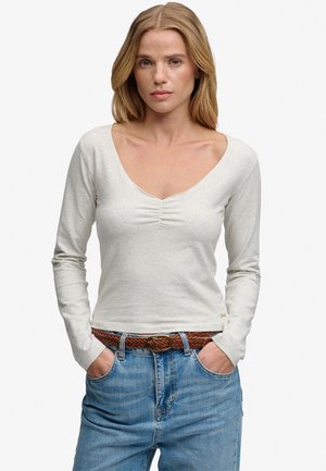 Kobieta o blond włosach, ubrana w jasnoszary top z długim rękawem i marszczonym dekoltem, niebieskie dżinsy i brązowy pleciony pasek, z rękami w kieszeniach.