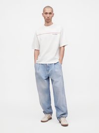 Homme debout les mains dans les poches, portant un t-shirt blanc à manches courtes, un jean large bleu clair et des baskets blanches sur un fond uni.