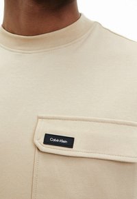 Beżowa bawełniana koszulka z wysokim kołnierzem, z kieszonką na klatce piersiowej i czarną metką Calvin Klein. Gładka faktura, prosty design.