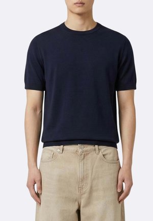 Uomo che indossa una maglietta blu scuro tinta unita a maniche corte con scollo rotondo e pantaloni beige chiaro, in piedi davanti a uno sfondo chiaro uniforme.