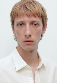 Weißes Hemd mit klassischen Kragen, das eine glatte Textur aufweist. Das Modell hat hellblonde Haare und blaue Augen, vor einem neutralen Hintergrund.