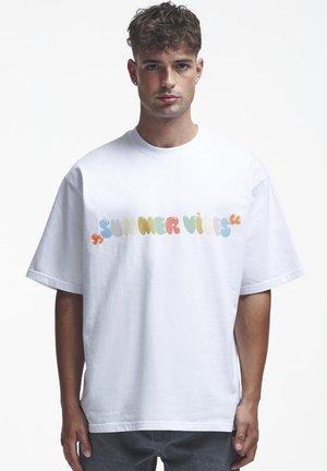 Fiatal férfi rövid göndör hajjal, fehér, bővebb, színes "SUMMER VIBES" feliratú pólóban, a mellkasán.