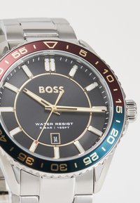 Ασημένιο ρολόι από ανοξείδωτο ατσάλι με μαύρο καντράν, χρυσές λεπτομέρειες, αδιάβροχο, bezel σε μπορντό και πράσινο χρώμα, και ένδειξη ημερομηνίας στις 6 η ώρα.