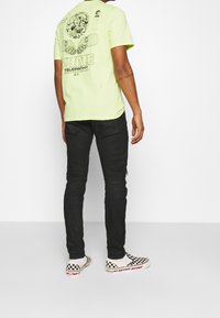 Person som står vänd bort med ansiktet, iklädd en neongul grafisk t-shirt, svarta slim fit jeans och svart-vita rutiga slip-on skor.