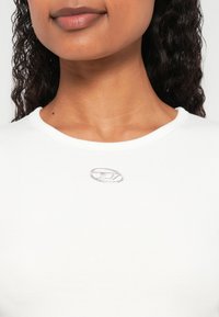 Fehér bordázott póló kerek nyakkivágással és egy ezüst ovális logó díszítéssel a mellrész fölött. Sima textúra, testhezálló szabás.