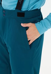 Hand ruht in der Tasche von dunkeltürkisfarbenen Outdoor-Hosen mit verstellbarem Bund, der Manschette eines blauen Langarmshirts sichtbar.