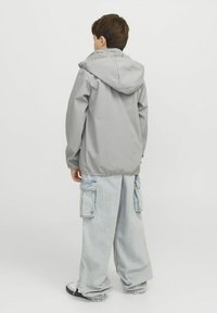 Grijze hooded jacket van synthetisch materiaal, gedragen met lichtblauwe ruime cargobroeken. Heeft elastische manchetten en een ontspannen pasvorm.