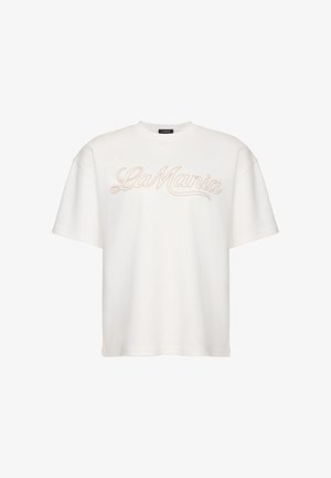 Tricou alb oversized cu mâneci scurte, cu „La Mania” brodat în bej scris cursiv pe piept, guler rotund și o etichetă neagră în interiorul gulerului.