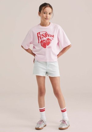 Noor tüdruk seisab käed puusal, seljas roosa "Maasikafestivali" T-särk, hele teksapüksid, valged sokid punaste triipudega ja hõbedased tossud.