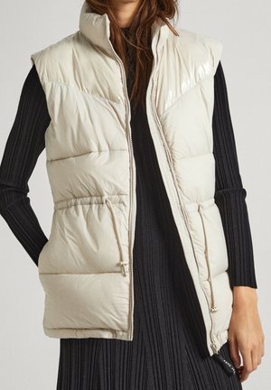 Femme portant une veste sans manches matelassée beige sur une robe noire en maille côtelée à manches longues, les mains dans les poches de la veste.