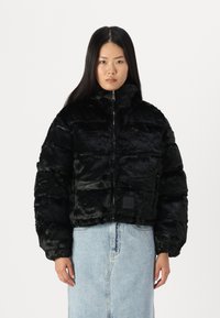 Jachetă puffer cropped din blană faux neagră, cu secțiuni matlasate, guler înalt și închidere cu fermoar. Include o mică etichetă cu marcă pe față.