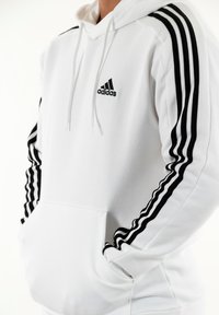 Sudadera blanca con el logo negro de Adidas y tres franjas negras en las mangas. Cuenta con un bolsillo frontal tipo pouch y capucha con cordón.