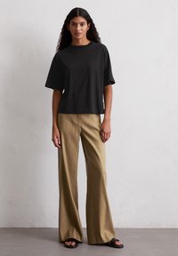T-shirt nero a maniche corte, aderente e dalla vestibilità ampia, abbinato a pantaloni khaki larghi e a vita alta e sandali neri su uno sfondo neutro.