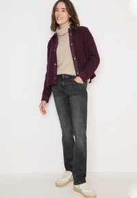 Chaqueta burdeos texturizada con botones lilas sobre un cuello de tortuga beige, combinada con jeans negros y zapatillas blancas con acentos azules.
