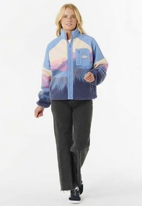 Veste en polaire bleue et rose avec fermeture éclair à l'avant, présentant un motif de montagne et une poche poitrine. Associée à un jean foncé et des baskets.