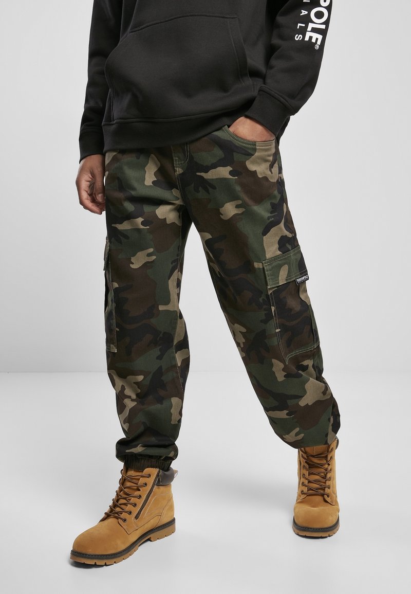 Camouflage cargo pants in green, brown, and black hues, featuring side pockets and a relaxed fit, paired with tan lace-up boots.