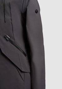 Temno siva rokav jakne z diagonalnim žepom z zadrgo in črnim gumbom na zgornjem delu roke na enostavni svetli podlagi.