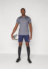 Tricou sport gri, pantaloni scurți bleumarin, șosete de fotbal negre și ghete de fotbal. Modelul ține o minge de fotbal alb-negru cu model. Design simplu, aspect ajustat.