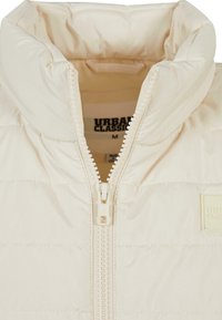 Beige Pufferjacke mit hohem Kragen, gerippter Textur, Frontreißverschluss und einem Logoelement auf der linken Seite. Leichtes, gestepptes Design.