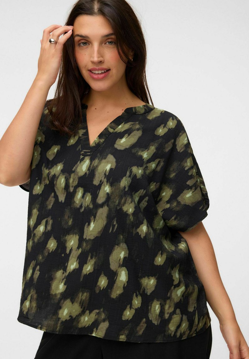 Femme aux longs cheveux foncés vêtue d'un chemisier noir ample avec un motif abstrait vert et des manches courtes, posant avec la main près du visage.