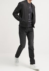 Musta bomber-takki, jolla on sileä pinta, puettuna tumman graafisen t-paidan päälle, yhdistettynä haalistuneisiin mustiin farkkuihin ja valkoisiin tennareihin.