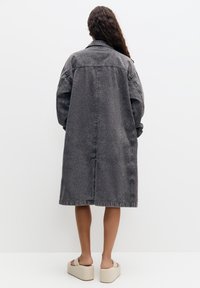 Ylipitkä harmaa farkkutakki, jossa on kaulus, roikkuvat olkap äät ja suora siluetti. Takissa on sauma-yksityiskohtia ja polvimittainen leikkaus.