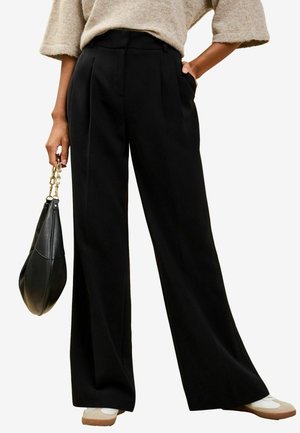 Weite schwarze Hose mit vorderen Falten, hoher Taille und Seitentaschen. Kombiniert mit einem lässigen Pullover und hält eine schwarze Handtasche.