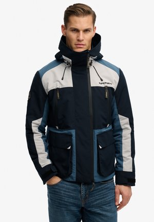 Homme portant une veste à capuche bleue, marine et grise avec poches avant et fermeture éclair, debout avec une main dans la poche sur fond blanc.