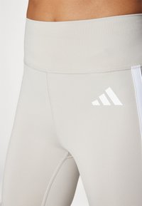 Lys grå treningstights med høy midje, prydet med hvite sidestriper og en liten hvit logo. Glatt, strammende stoff.