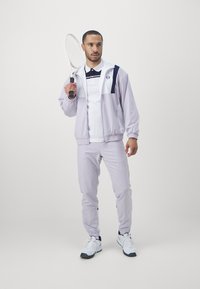 Helllila Trainingsanzug mit einem weißen und marineblauen Poloshirt. Die Jacke hat einen Reißverschluss und gerippte Bündchen. Das Model hält einen Tennisschläger. Weiße Sneakers.