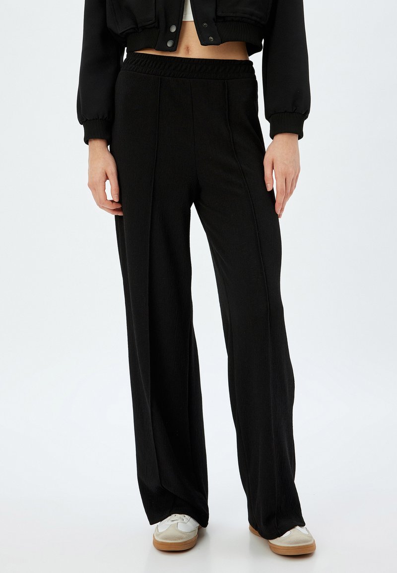 Pantalones amplios negros con una tela texturizada, cintura elástica y detalles de costura central, combinados con zapatillas blancas que tienen acentos claros.