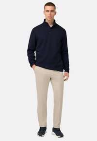 Jūras spēku pārpulloveris ar augstu apkakli, trim pogām un teksturētu adījumu, apvienots ar gaismas bēšiem biksēm un tumšiem sporta apaviem.