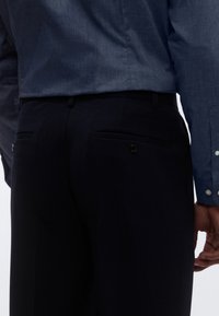 Tamsiai mėlynos spalvos kelnės su pritaikytu pjūviu, turinčios du kišenes gale su sagomis, derinamos su tekstūruota mėlyna marškinių palaidine su sagomis.