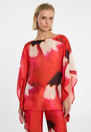 Femme portant un chemisier ample tie-dye rouge, rose et noir avec des manches larges et un pantalon assorti, sur un fond blanc uni.