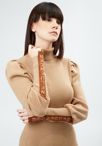 Pull en tricot beige avec un col montant, des épaules bouffantes et des poignets contrastants marron avec du texte. Texture douce et silhouette ajustée.