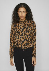 Blouse à imprimé léopard marron et noir avec des manches longues, un col froncé et des poignets élastiques, assortie à un pantalon noir ajusté.