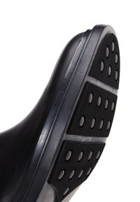 Suola di scarpa in gomma nera con superficie texturizzata, caratterizzata da motivi di presa circolari e un bordo liscio. Leggera curvatura nel tallone.