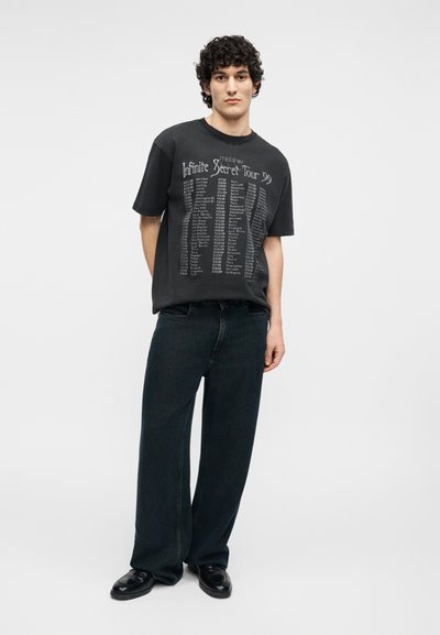 Νεαρός με σγουρά μαλλιά φορά μαύρο T-shirt με την επιγραφή «Infinite Secret Tour '99», φαρδιά σκούρα τζιν και μαύρα παπούτσια, στέκεται μπροστά σε απλό φόντο.