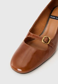 Brown leather shoe with a rounded toe, cut-out detail, and a front strap secured with a gold-tone buckle. Smooth, shiny finish.