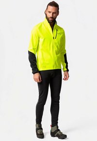 Rumena jakna z visoko vidnostjo z črnimi rokavi, z zadrgo spredaj in odsevnimi detajli. Obuta čez črne športne pajkice in čevlje.