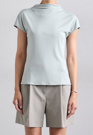 Kvinde iført lyseblå top med korte ærmer, slået krave og højtaljede beige shorts, stående foran en ensfarvet grå baggrund.