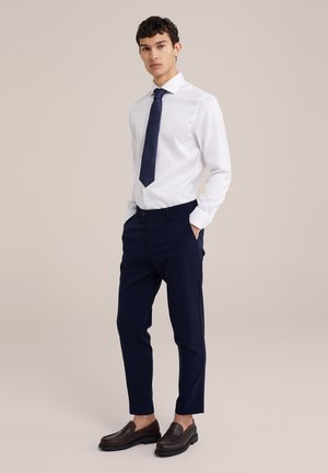 Camicia bianca, pantaloni blu su misura, cravatta scura a fantasia, mocassini marroni. Vestibilità slim, tessuto morbido e dettagli di cucitura accentuati.