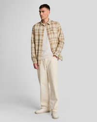 Beige og brunternet skjorte med lange ærmer og knapper, båret over en lys t-shirt, kombineret med cremefarvede bukser med vide ben.