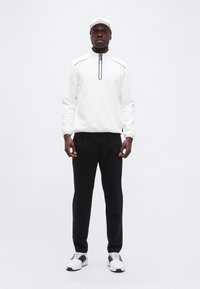 Ανδρας με λευκό φούτερ με φερμουάρ έως τη μέση, λευκό καπέλο, μαύρο παντελόνι και λευκά αθλητικά παπούτσια, που στέκεται μπροστά σε απλό φωτεινό φόντο.