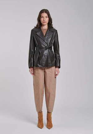 Kobieta w czarnej skórzanej kurtce z paskiem, beżowych spodniach i brązowych botkach do kostki, stojąca na tle jednolitego jasnoszarego tła.