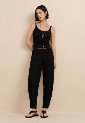 Vrouw staat tegen een beige muur, draagt een zwart tanktopje, losse zwarte broek met gouden accenten, zwarte sandalen en een ketting met een sterhanger.