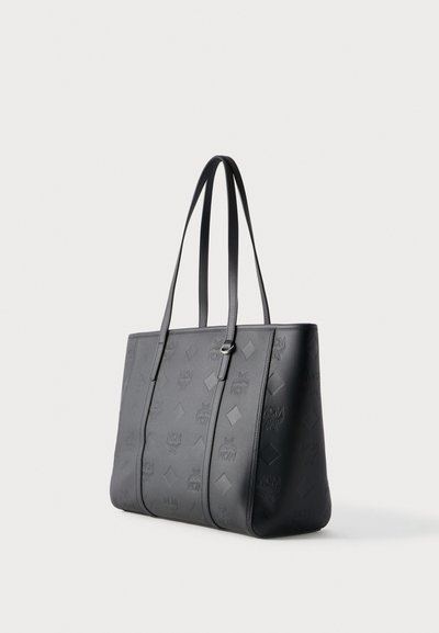 Sac cabas en cuir noir avec logos MCM embossés et longues bretelles doubles sur fond blanc.